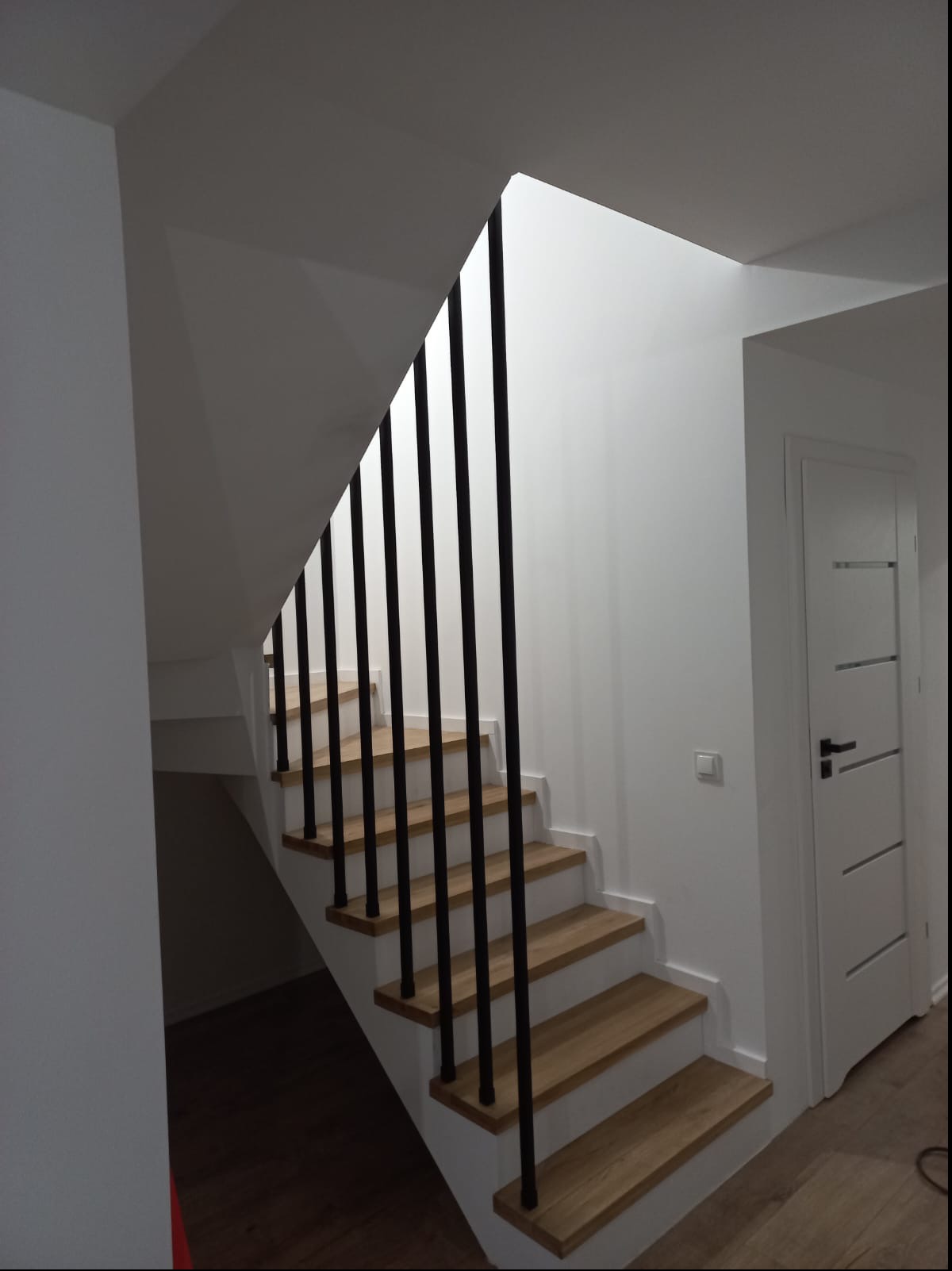 Klatka schodowa z drewnianymi stopniami i czarną, minimalistyczną balustradą pionową, oświetlona naturalnym światłem w nowoczesnym wnętrzu.