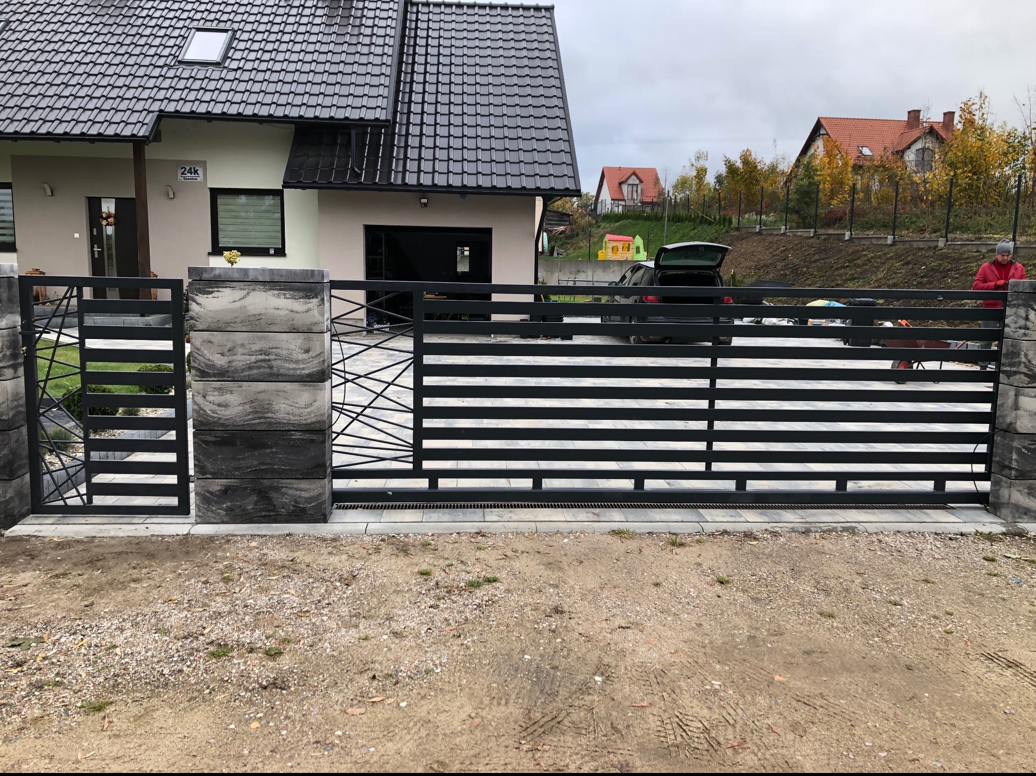 Nowoczesna, przesuwna brama wjazdowa z czarnymi, poziomymi panelami i betonowymi słupkami, widok z zewnątrz posesji, dom jednorodzinny w tle.