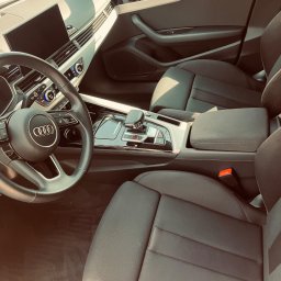 Maciej Wyżga - Wnętrze samochodu Audi A4 z czarną tapicerką, widoczna kierownica z logo Audi, deska rozdzielcza z ekranem i automatyczna skrzynia biegów.