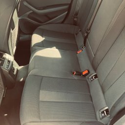 Maciej Wyżga - Wnętrze samochodu: tylna kanapa z szarej tapicerki z widocznymi pasami bezpieczeństwa i kratką na drobiazgi na oparciu przedniego fotela.