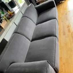 Szara, modułowa sofa z widocznymi szwami na tle jasnej podłogi z drewna i okna z widokiem na zieleń.