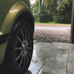 Maciej Wyżga - Fragment umytego, zielonego samochodu z aluminiowymi felgami na mokrych, szarych płytkach chodnikowych, widok z niskiej perspektywy.