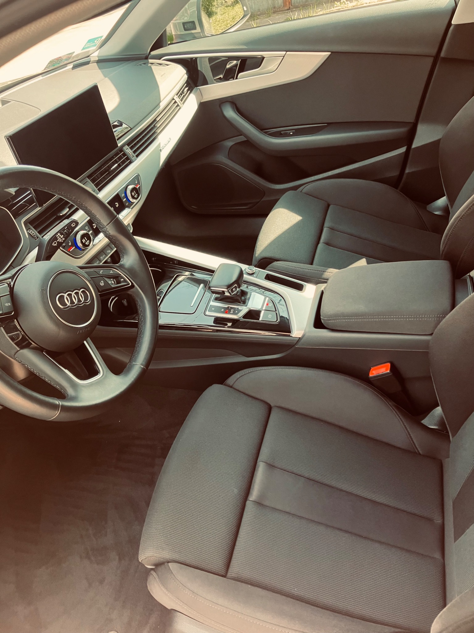 Wnętrze samochodu Audi A4 z czarną tapicerką, widoczna kierownica z logo Audi, deska rozdzielcza z ekranem i automatyczna skrzynia biegów.