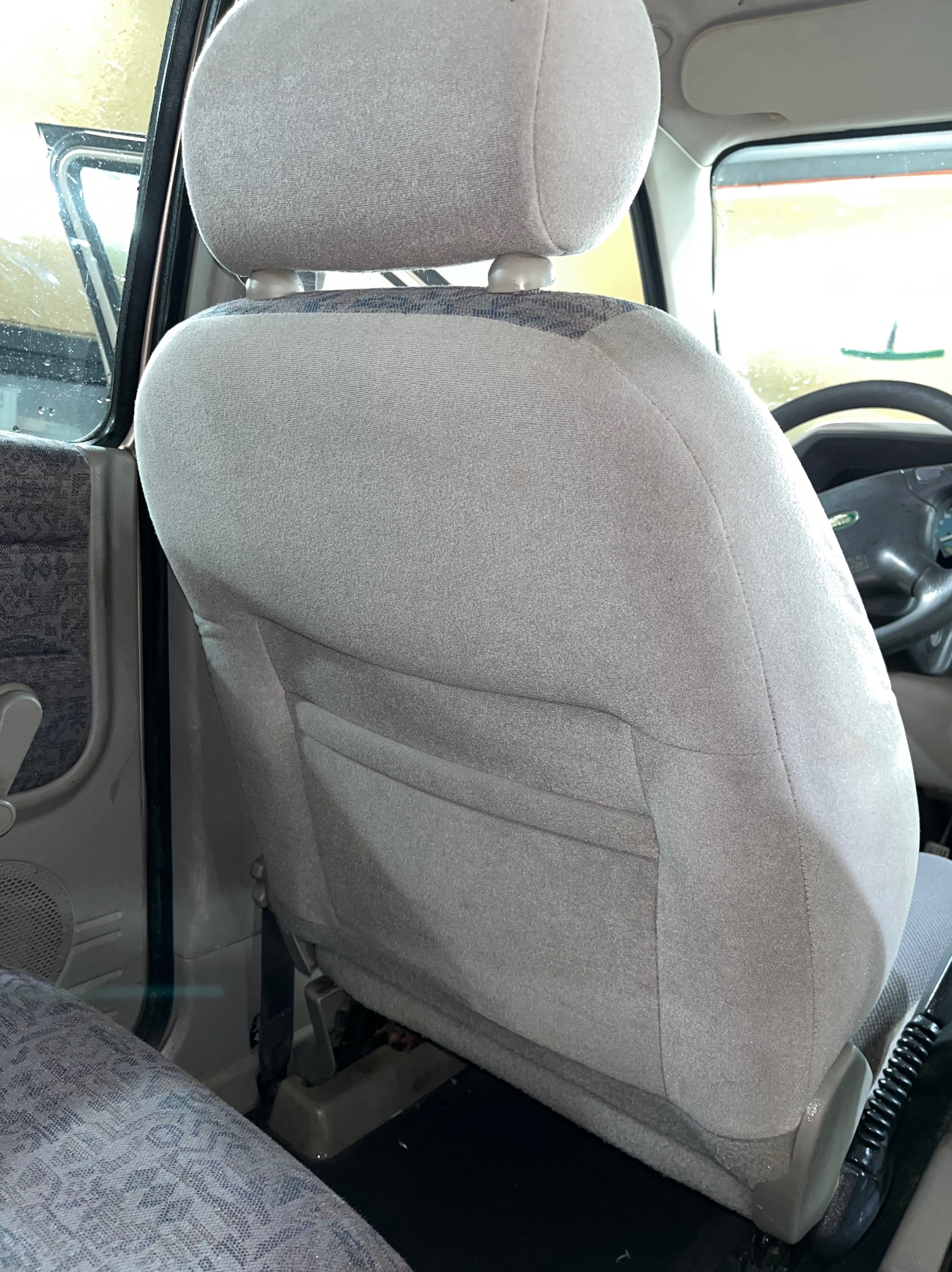 Szare, materiałowe siedzenie samochodowe z widocznymi plamami i zabrudzeniami, od strony oparcia, z kieszenią z tyłu.