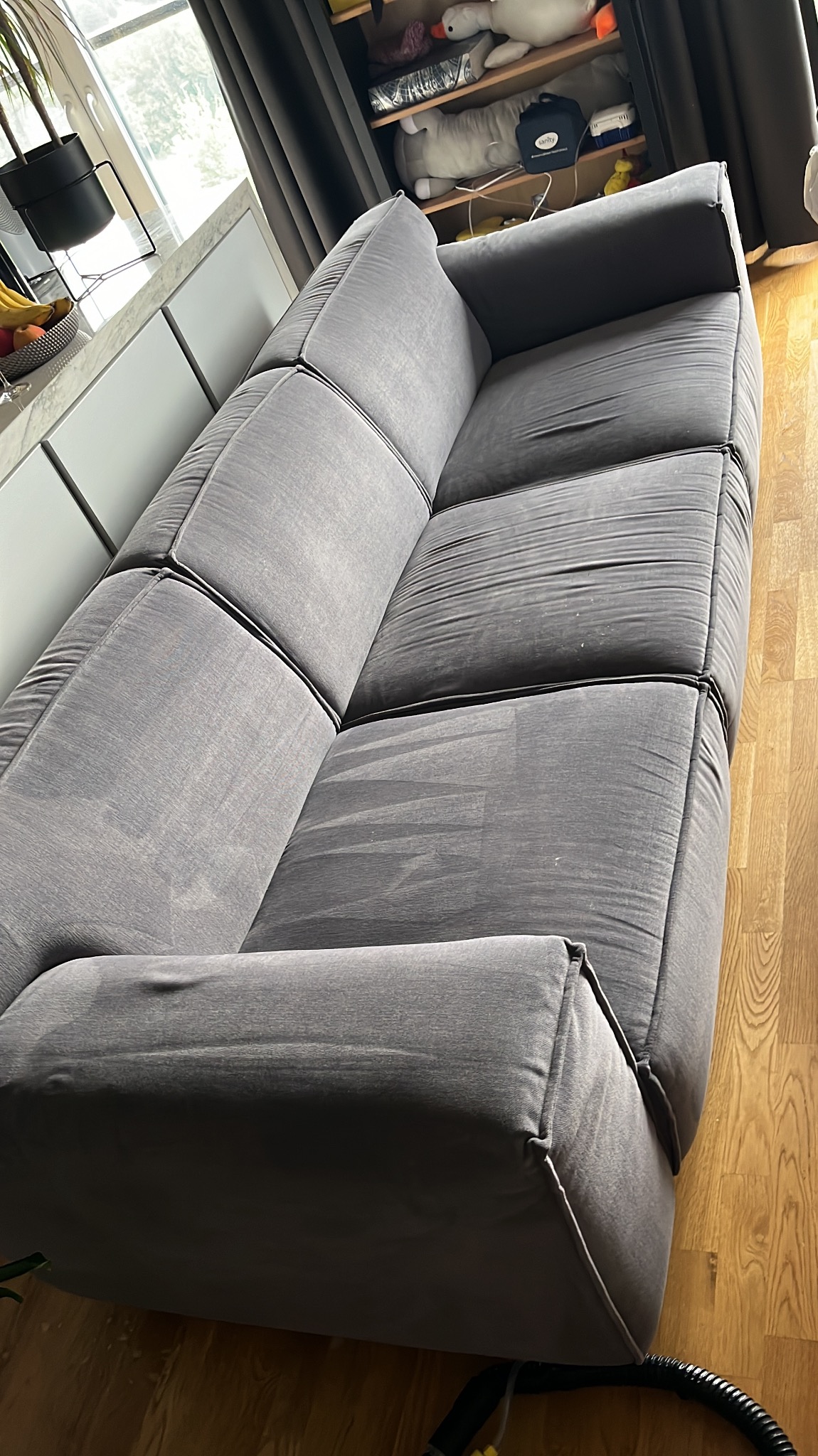 Szara, modułowa kanapa z widocznymi śladami po praniu tapicerki, ukazującymi różnicę w czystości, na drewnianej podłodze, z fragmentem urządzenia do czyszczenia w dolnym rogu.