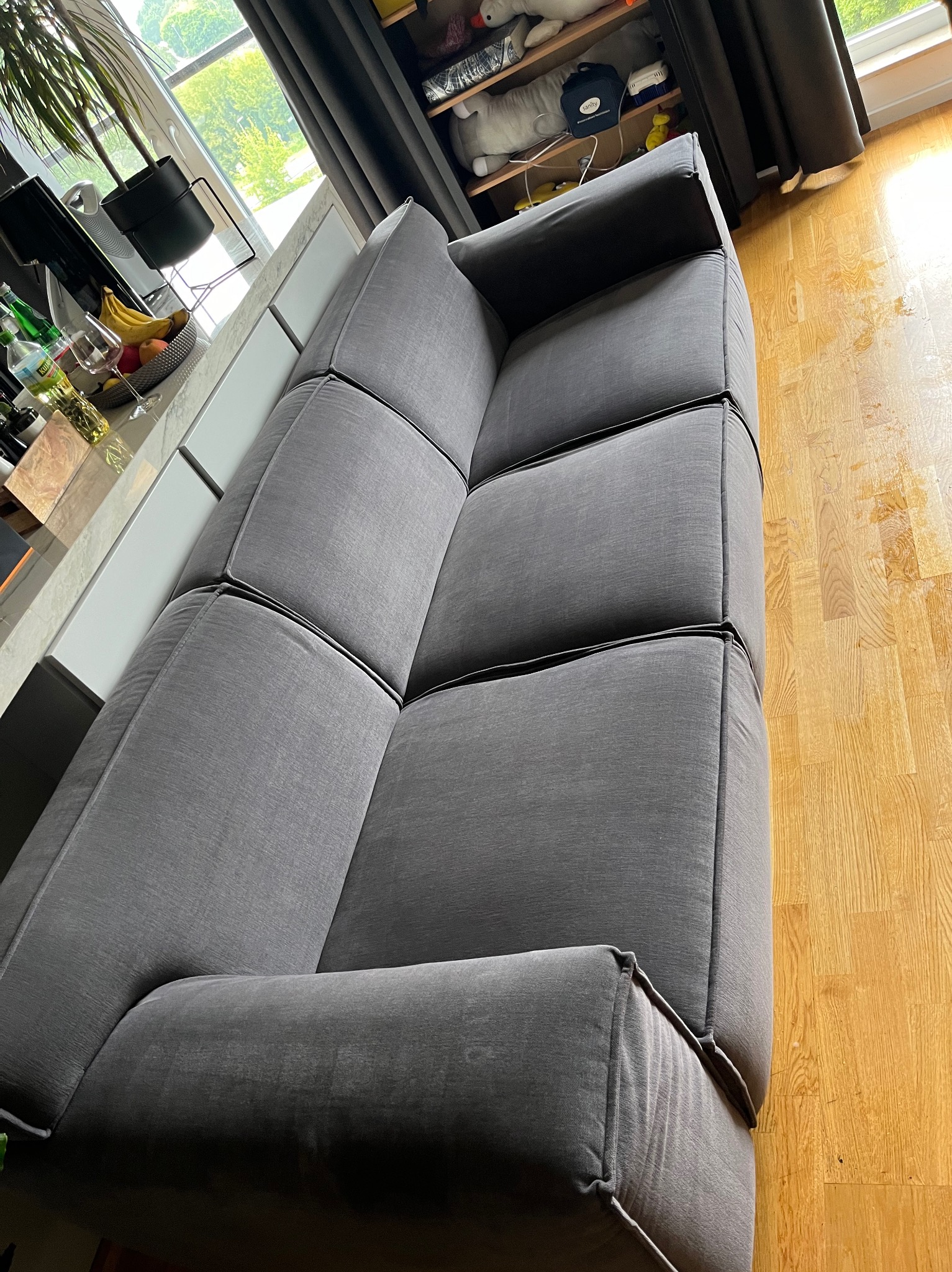 Szara, modułowa sofa z widocznymi szwami na tle jasnej podłogi z drewna i okna z widokiem na zieleń.