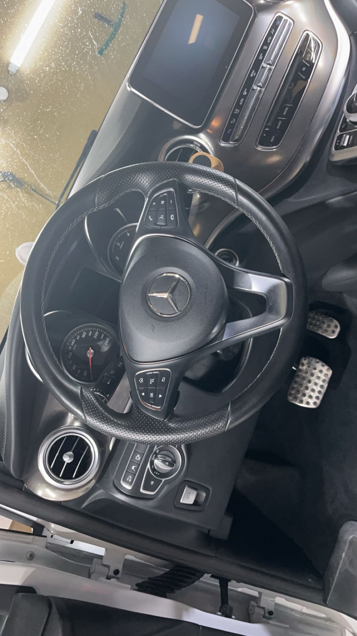 Wnętrze samochodu Mercedes, widok na kierownicę z logo, deskę rozdzielczą z ekranem i przyciskami, oraz metalowe pedały, po detailingu.