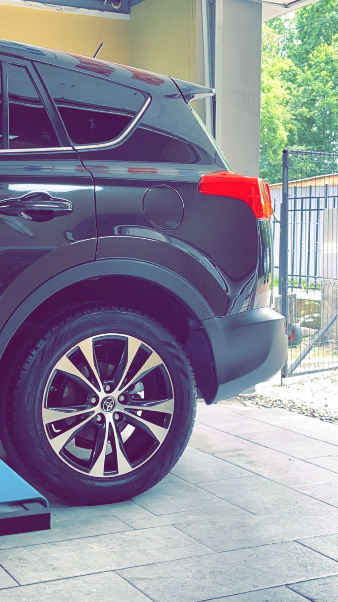 Część tylna czarnego samochodu Toyota RAV4 z widocznym kołem i felgą, zaparkowanego na jasnych płytkach chodnikowych, z zielonymi drzewami i metalowym ogrodzeniem w tle.