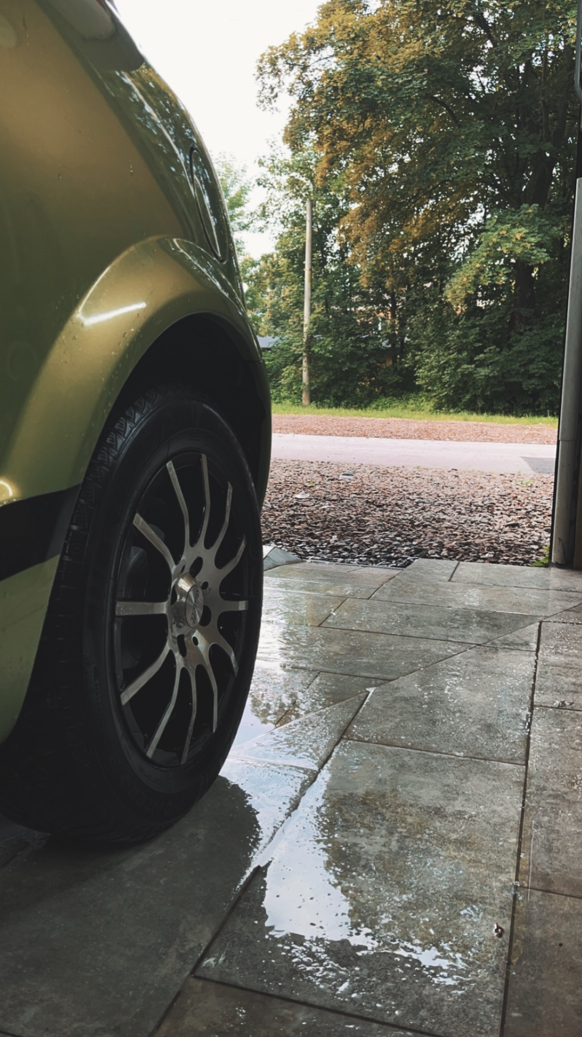 Fragment umytego, zielonego samochodu z aluminiowymi felgami na mokrych, szarych płytkach chodnikowych, widok z niskiej perspektywy.