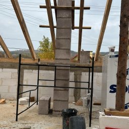 J3crewbau - Wznoszenie komina z bloczków betonowych na budowie domu jednorodzinnego, widoczne rusztowanie, drewniana więźba dachowa i materiały budowlane.