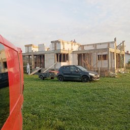 J3crewbau - Budowa domu z pustaków: ściany wzniesione, drewniane szalunki okienne, na dachu robotnicy. Na trawie zaparkowany samochód z przyczepką, obok czerwony bus.