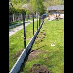 Montaż ogrodzenia panelowego: betonowa podmurówka z czarnymi słupkami na tle zieleni i błękitnego nieba. Widoczny wykop i mała koparka.