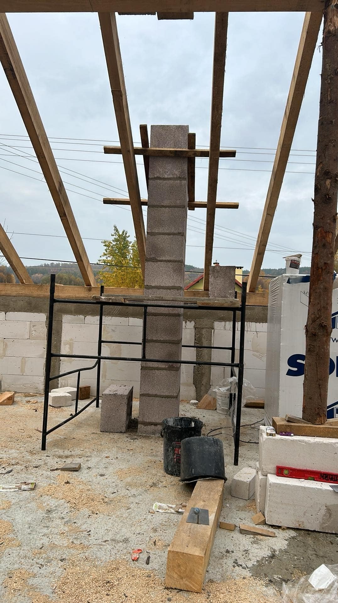 Wznoszenie komina z bloczków betonowych na budowie domu jednorodzinnego, widoczne rusztowanie, drewniana więźba dachowa i materiały budowlane.