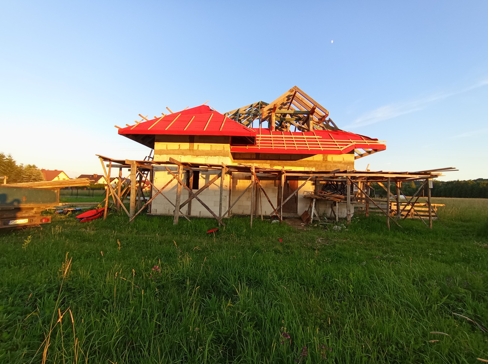 Niedokończony dom z czerwonym dachem i drewnianą konstrukcją, otoczony zielonym polem, w tle błękitne niebo z widocznym księżycem. Etap budowy.