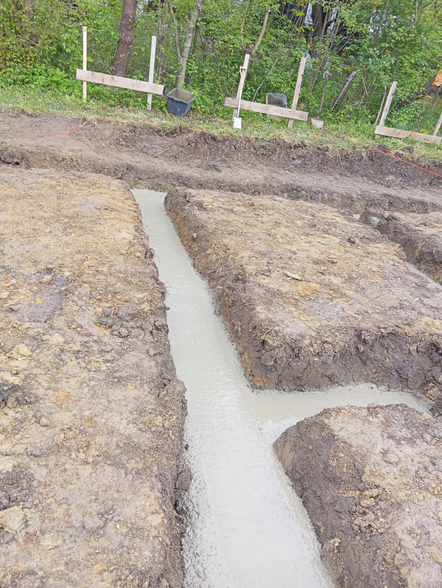Zalane betonem wykopy pod fundamenty z widocznymi deskami szalunkowymi i ziemią. Ujęcie z góry ukazuje etap wylewania fundamentów.