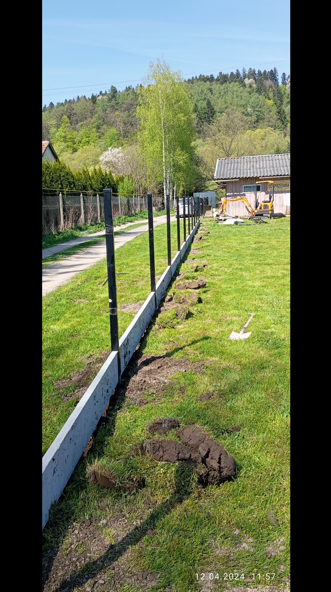 Montaż ogrodzenia panelowego: betonowa podmurówka z czarnymi słupkami na tle zieleni i błękitnego nieba. Widoczny wykop i mała koparka.