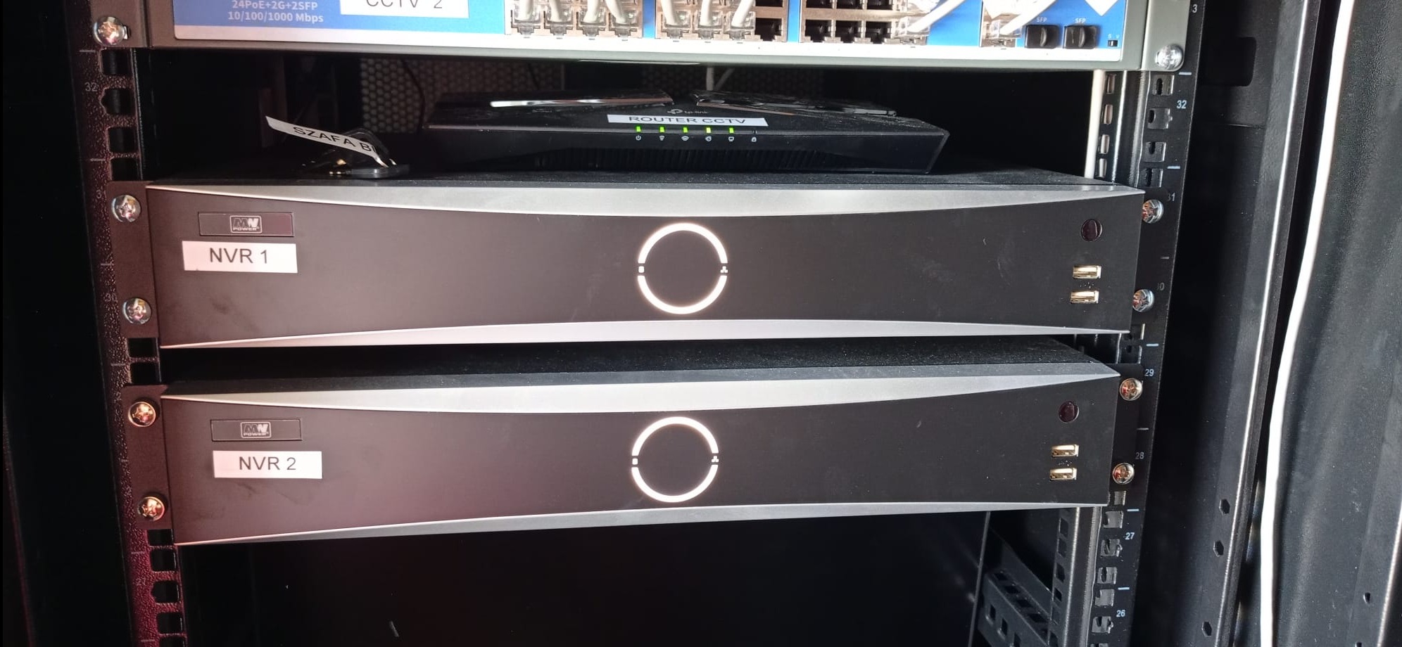 Dwa rejestratory NVR w szafie rack, z routerem CCTV na górze, widoczne oznaczenia 'NVR 1' i 'NVR 2'.