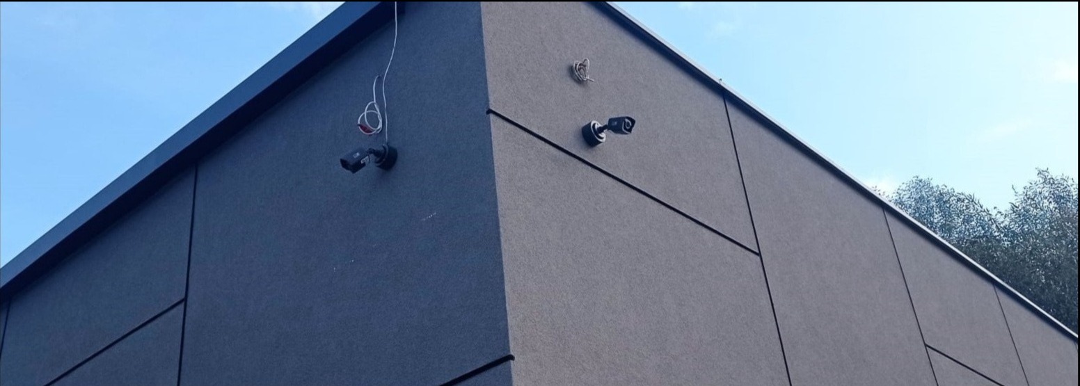 Dwie kamery monitoringu zamontowane na elewacji budynku o szarej, panelowej strukturze; jedna kamera z widocznymi przewodami, druga z zamaskowanym okablowaniem.