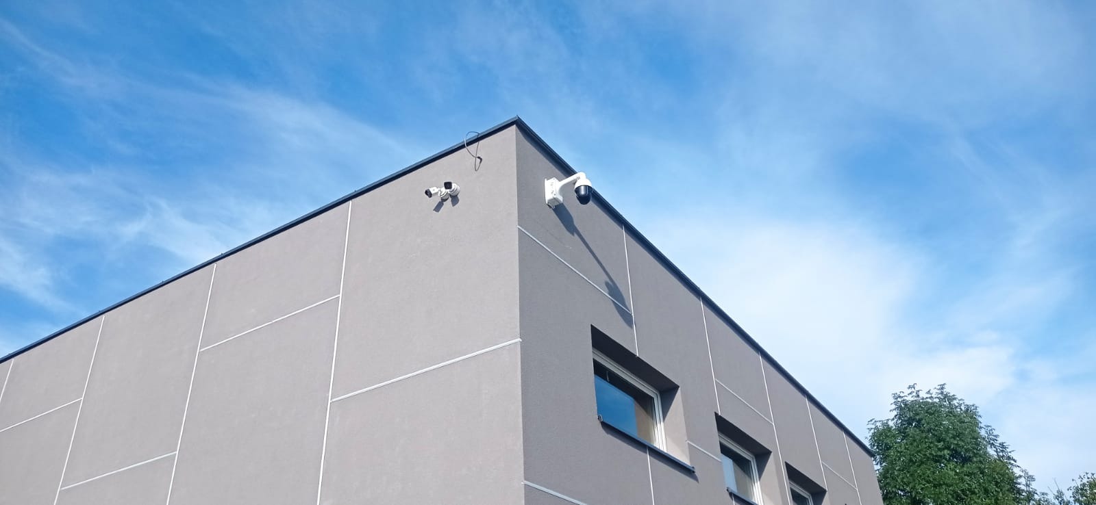 Dwie kamery monitoringu: jedna kopułkowa na wysięgniku, druga podwójna, zamontowane na szarym budynku z panelami, widok z dołu na błękitne niebo.