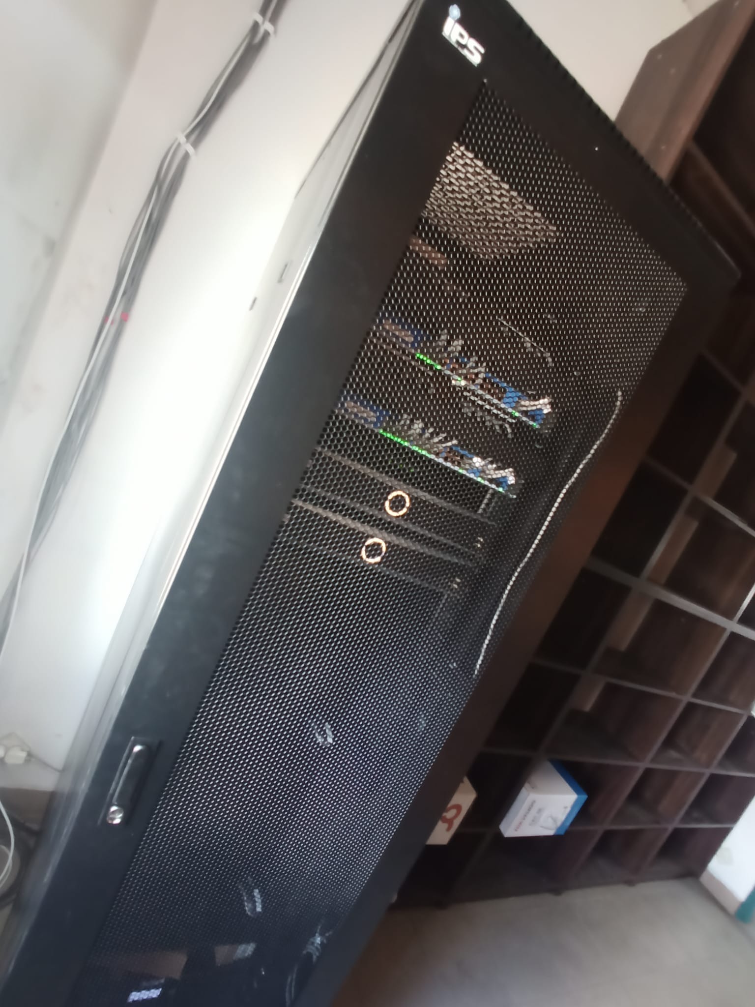 Czarna szafa rack z widocznymi kablami i urządzeniami wewnątrz, z logo IPS w górnym rogu, ustawiona w pomieszczeniu z półkami w tle.