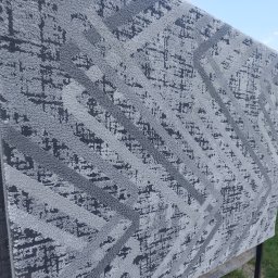 PLAMKO Paulina Pachnik - Szary dywan z geometrycznym wzorem w odcieniach szarości, suszony na zewnątrz na metalowej konstrukcji, widoczne fragmenty nieba i trawy.