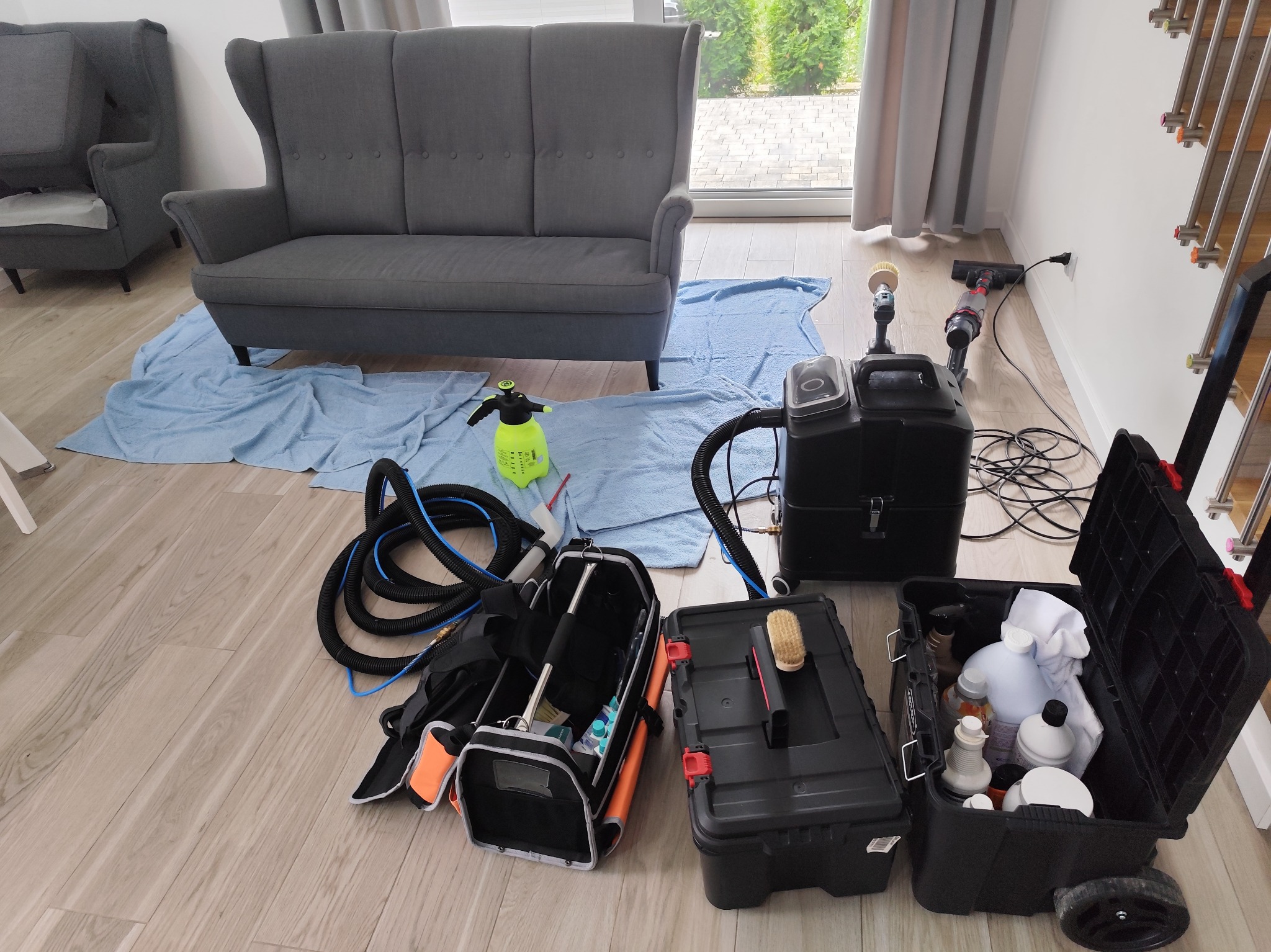 Przygotowanie do prania tapicerki: szara sofa zabezpieczona niebieskim ręcznikiem, obok rozłożone narzędzia i środki czystości w czarnych kufrach, widoczny spryskiwacz i odkurzacz piorący.