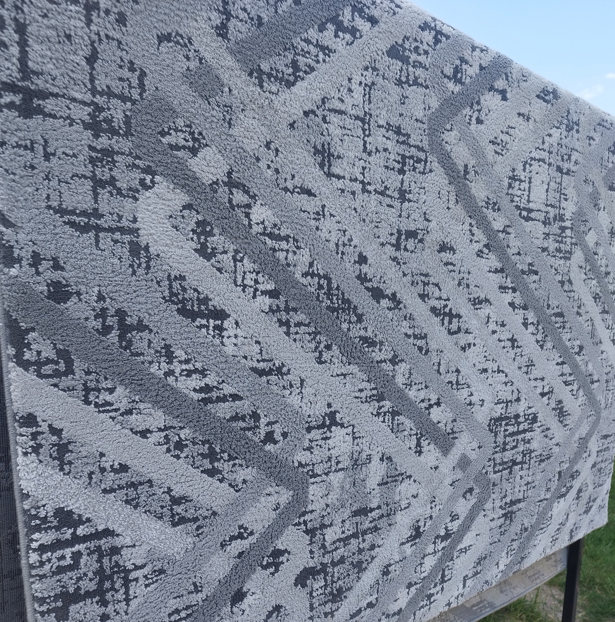 Szary dywan z geometrycznym wzorem w odcieniach szarości, suszony na zewnątrz na metalowej konstrukcji, widoczne fragmenty nieba i trawy.