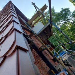 VR_BUD - Fragment dachu krytego dachówką karpiówką w odcieniach brązu, widoczny z dołu, z rusztowaniem w tle i fragmentem murowanej ściany szczytowej, częściowo zasłoniętej przez konstrukcję rusztowania.