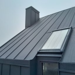 VR_BUD - Szary dach z blachy na rąbek stojący z oknem dachowym i kominem w deszczowy dzień.