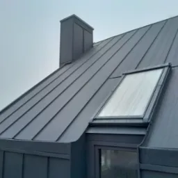 Szary dach z blachy na rąbek stojący z oknem dachowym i kominem w deszczowy dzień.