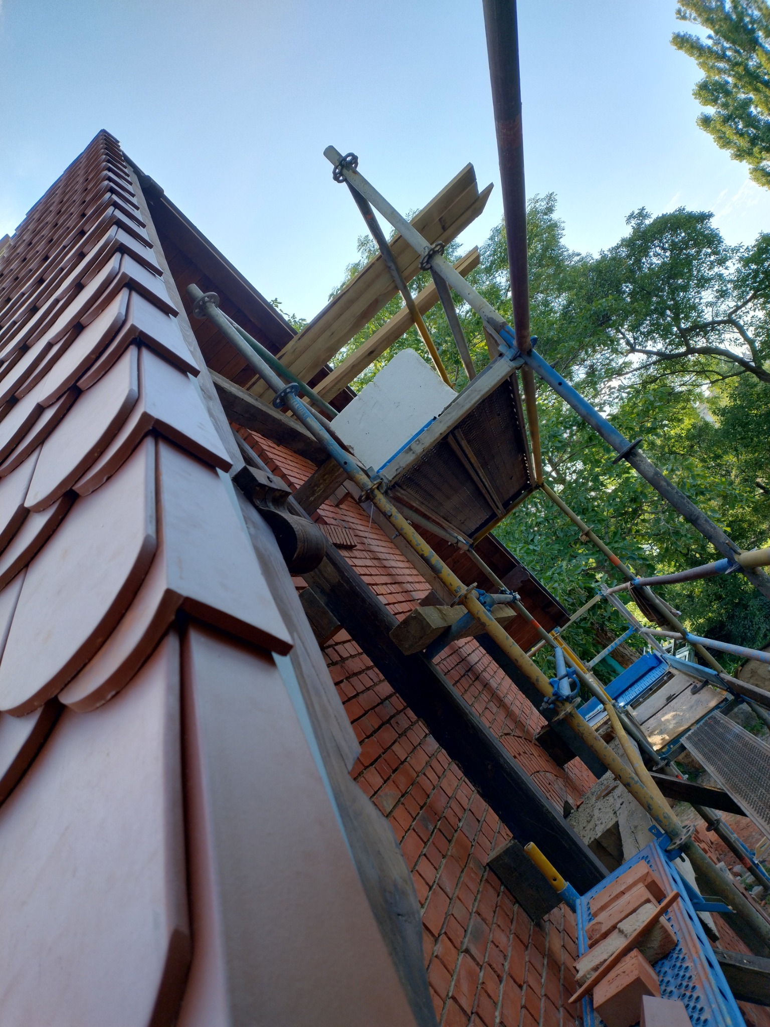 Fragment dachu krytego dachówką karpiówką w odcieniach brązu, widoczny z dołu, z rusztowaniem w tle i fragmentem murowanej ściany szczytowej, częściowo zasłoniętej przez konstrukcję rusztowania.
