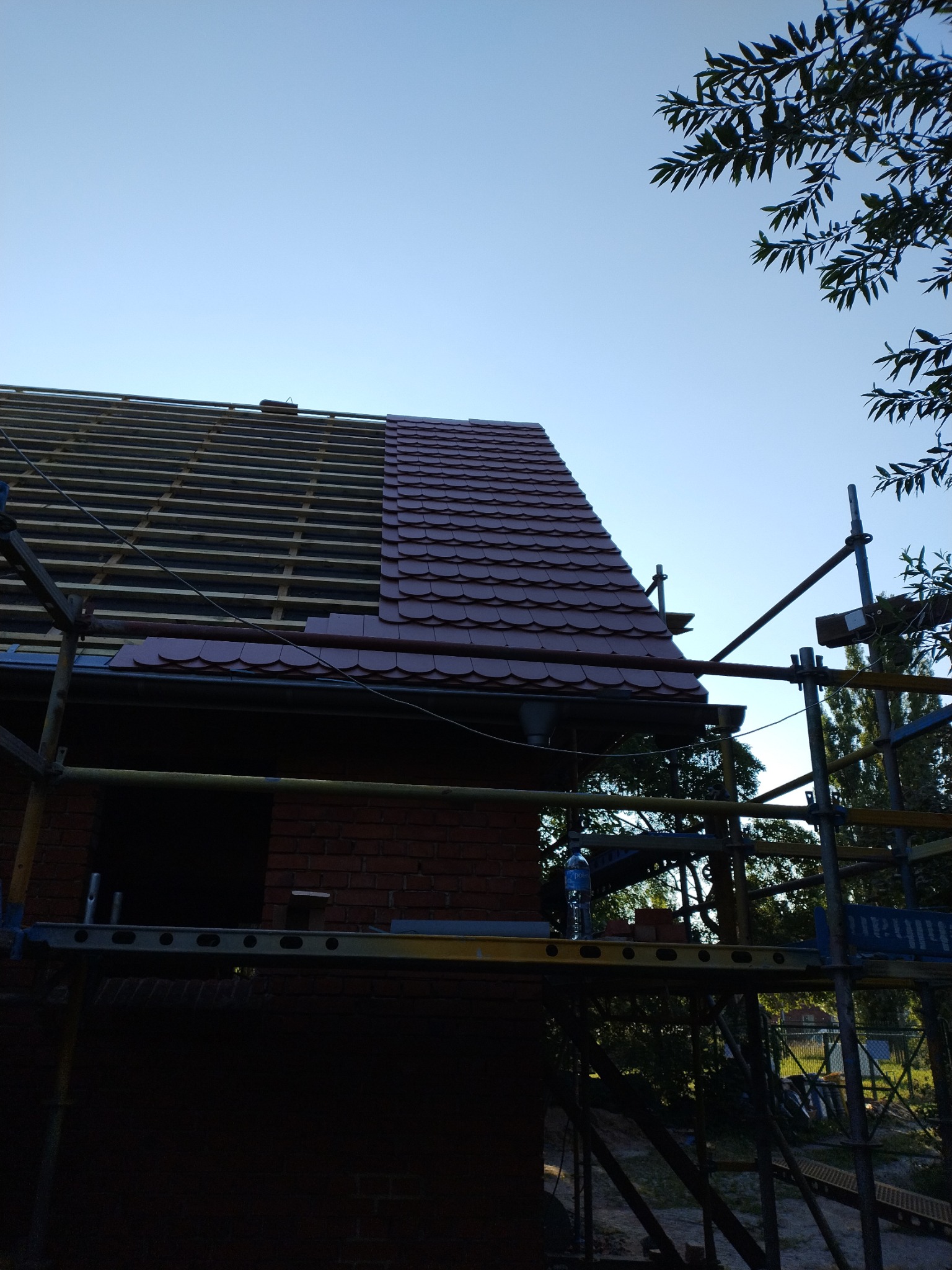 Częściowo pokryty czerwoną dachówką dach domu w Leźnie, widoczne rusztowanie i więźba dachowa.