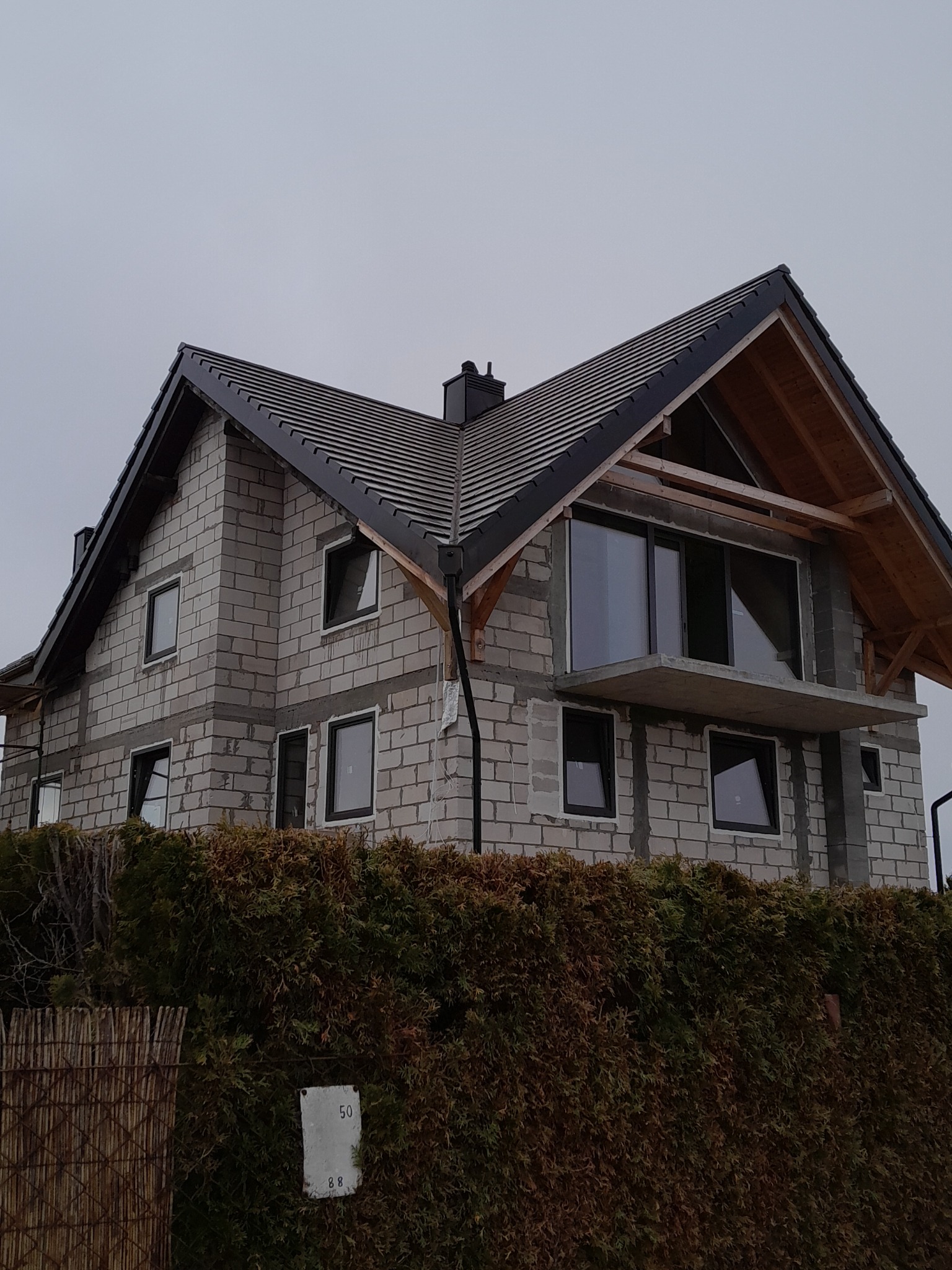 Dom jednorodzinny w trakcie budowy, z widocznym nowym dachem z ciemnej dachówki, surową elewacją z białych bloczków i częściowo zamontowanymi oknami, otoczony zielonym żywopłotem.