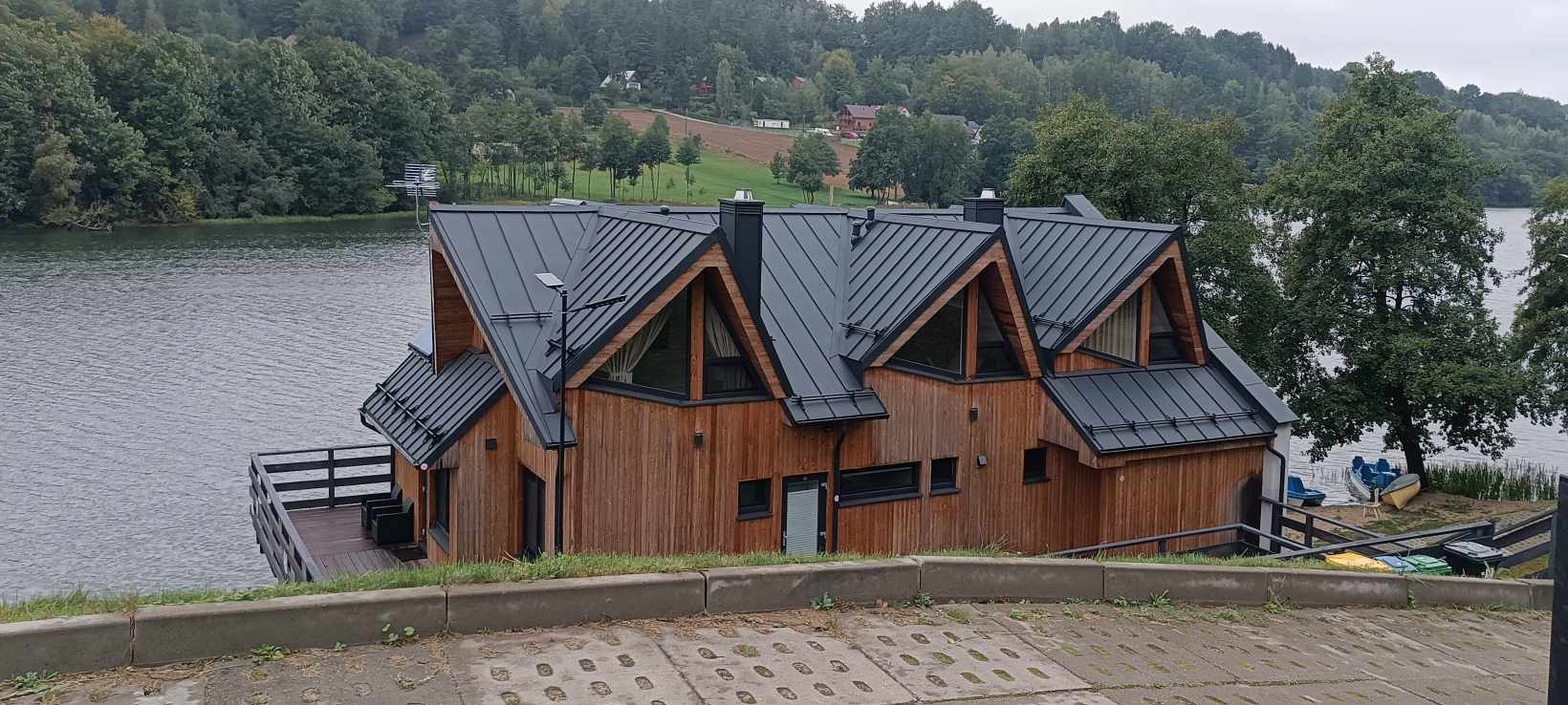 Drewniany dom z czarnym dachem z blachy na brzegu jeziora, widok z dołu na elewację i dach, z widocznym fragmentem tarasu i otaczającą zielenią.