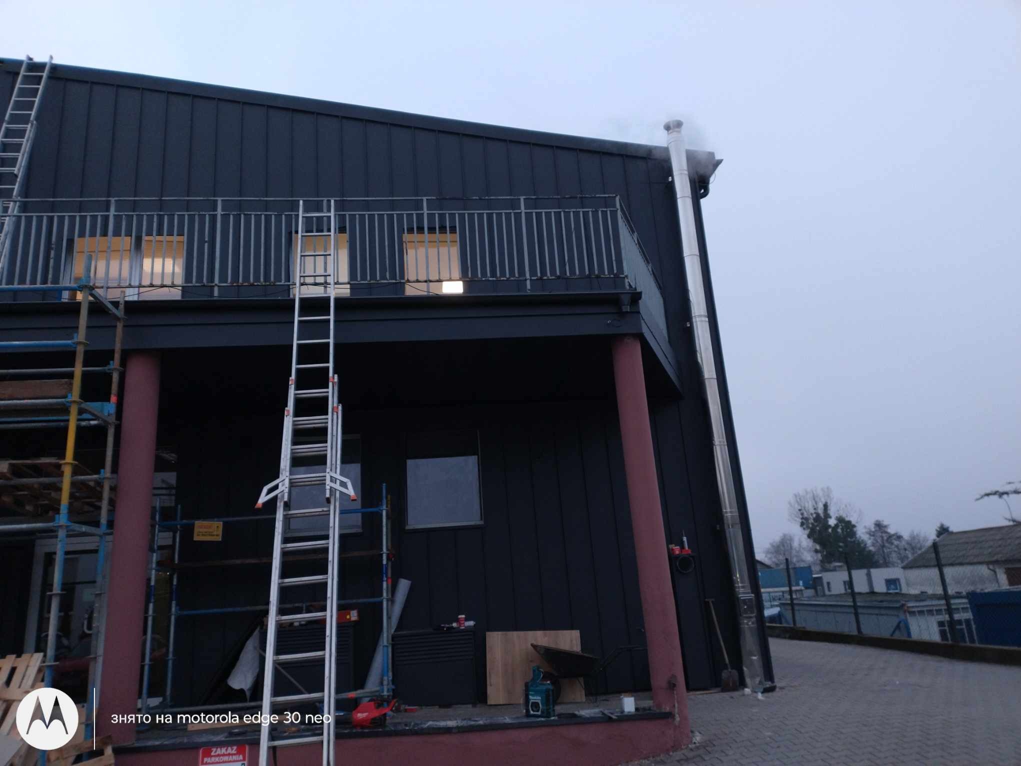 Budynek z czarną blachą na elewacji i dachu, widoczne rusztowania i drabiny, komin ze stali nierdzewnej.