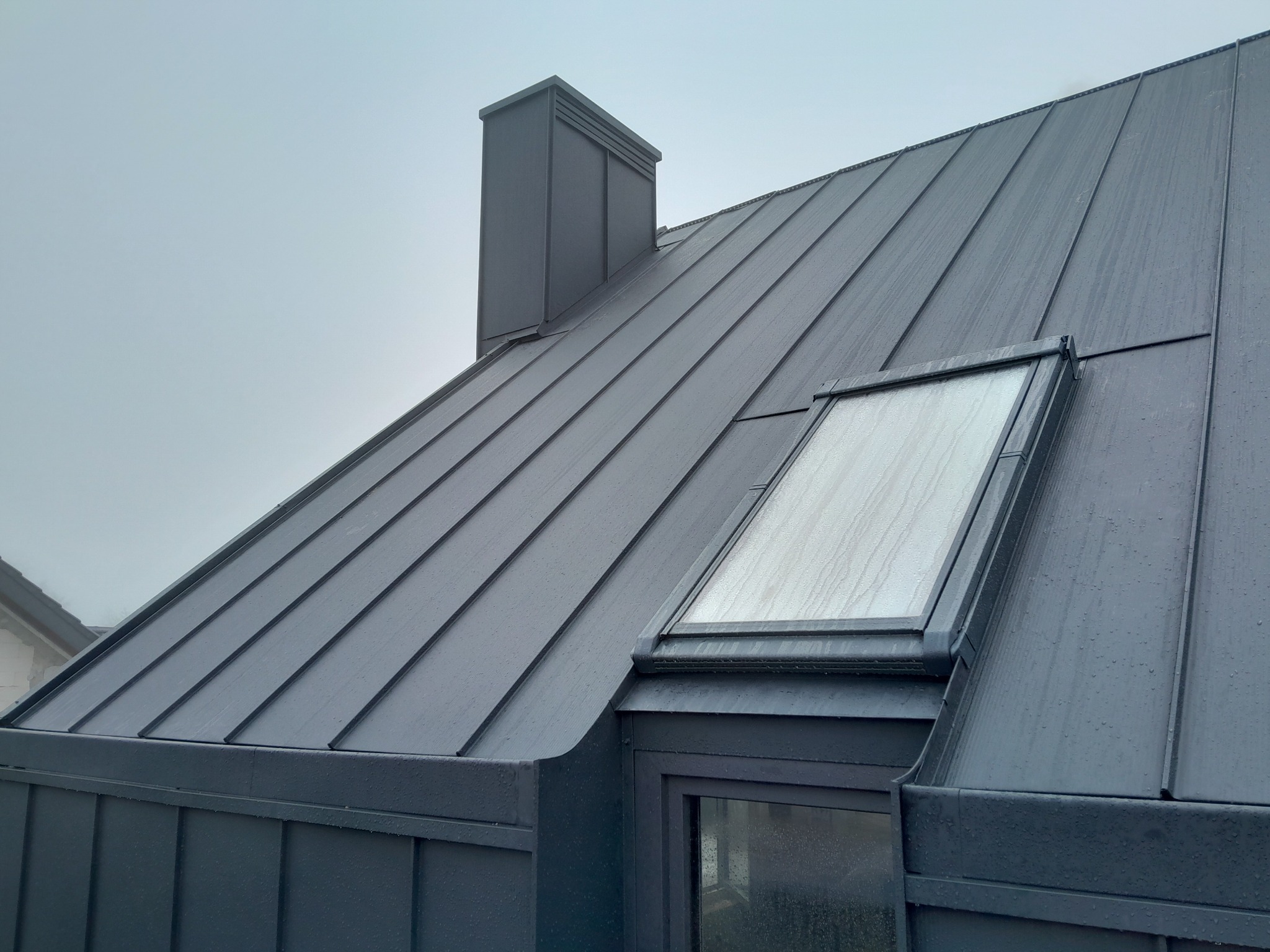 Szary dach z blachy na rąbek stojący z oknem dachowym i kominem w deszczowy dzień.