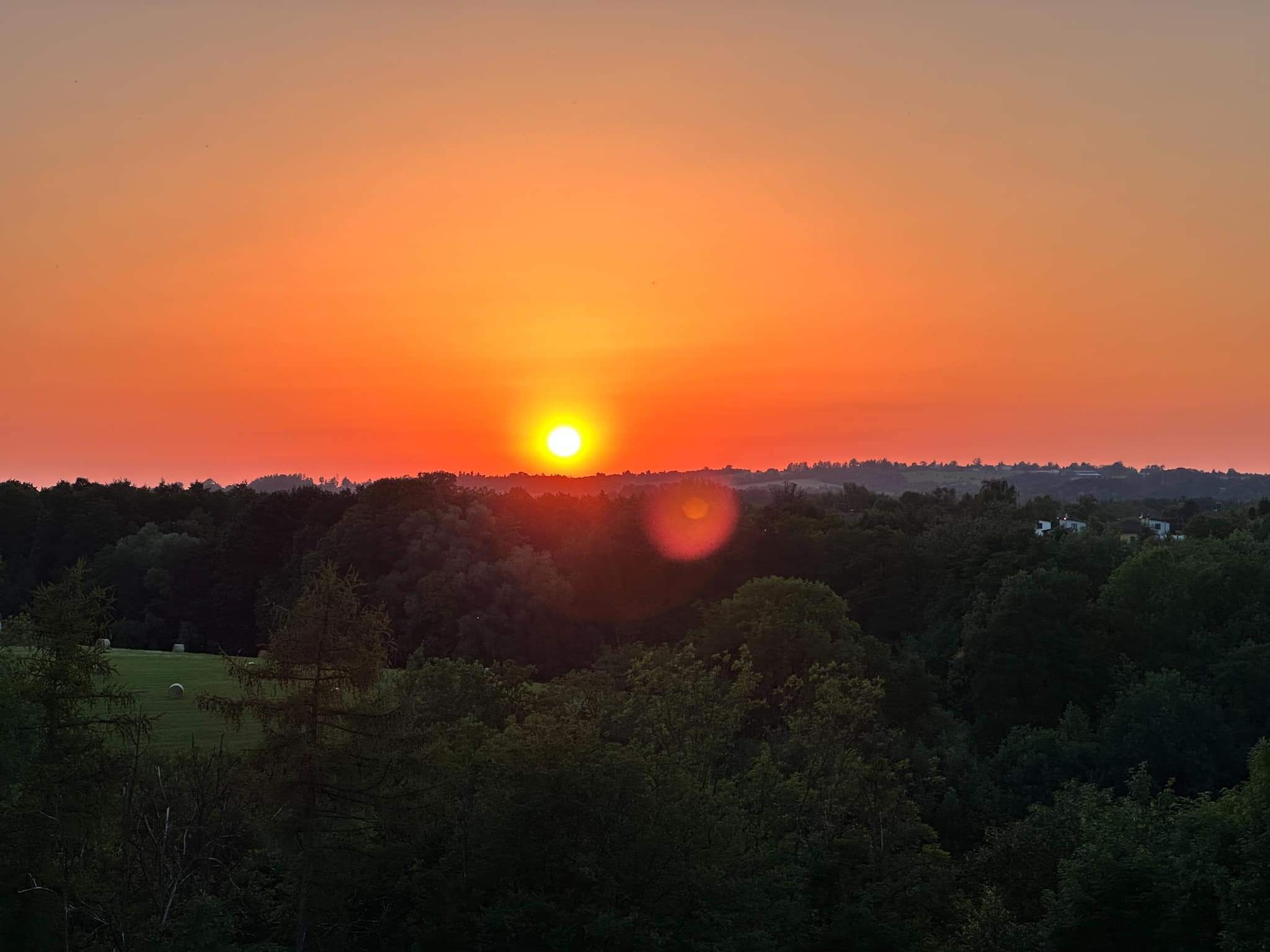 Pomarańczowe niebo o zachodzie słońca nad zalesionym terenem z belami siana na łące w oddali.
