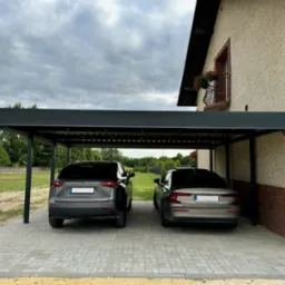 Nowoczesna wiata garażowa z płaskim dachem w kolorze antracytowym, kryjąca dwa samochody osobowe, z widoczną elewacją domu i balkonem z kwiatami w tle.