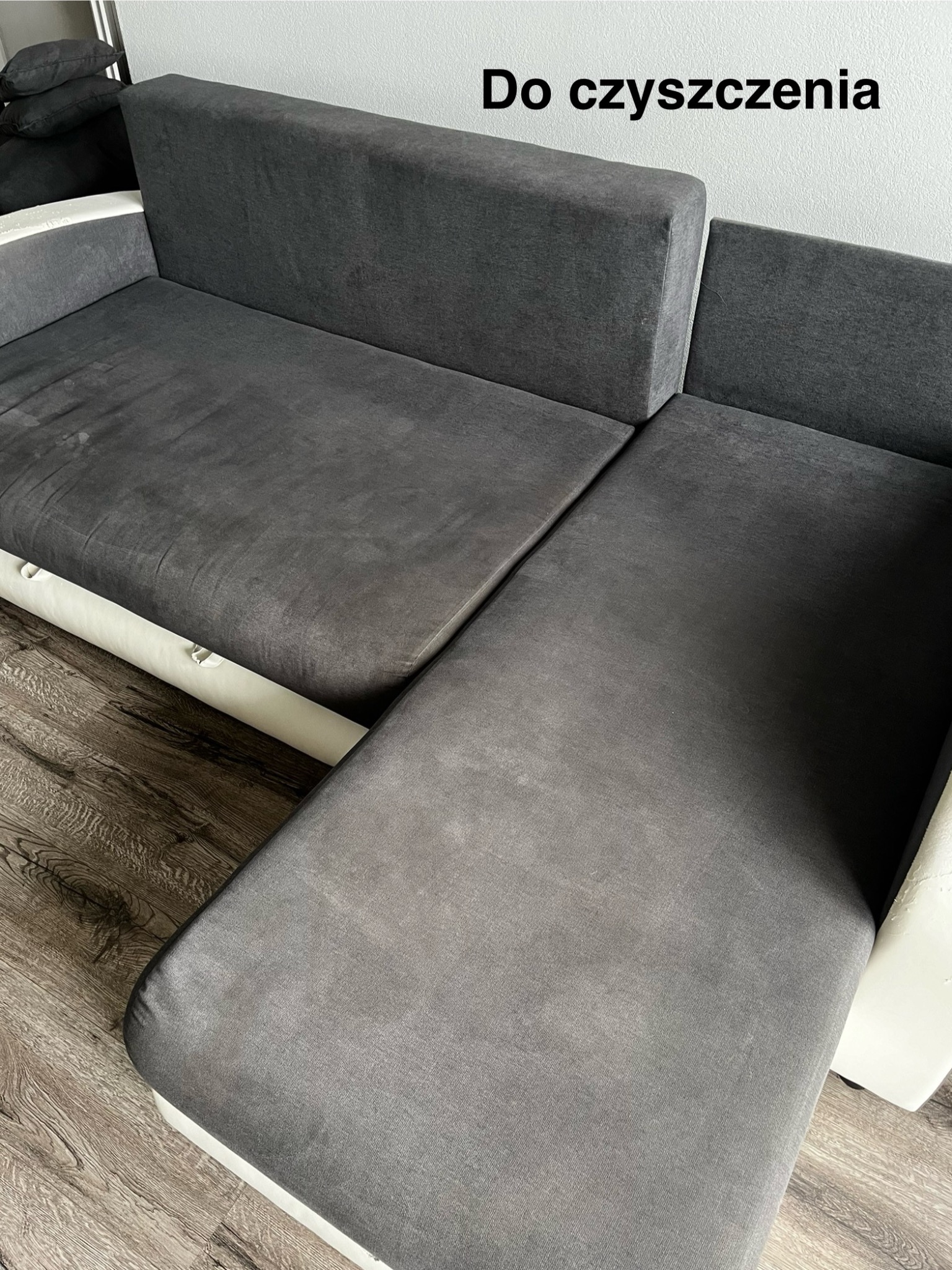 Szara, narożna kanapa z widocznymi plamami na tapicerce, wymagająca odświeżenia, na drewnianej podłodze.