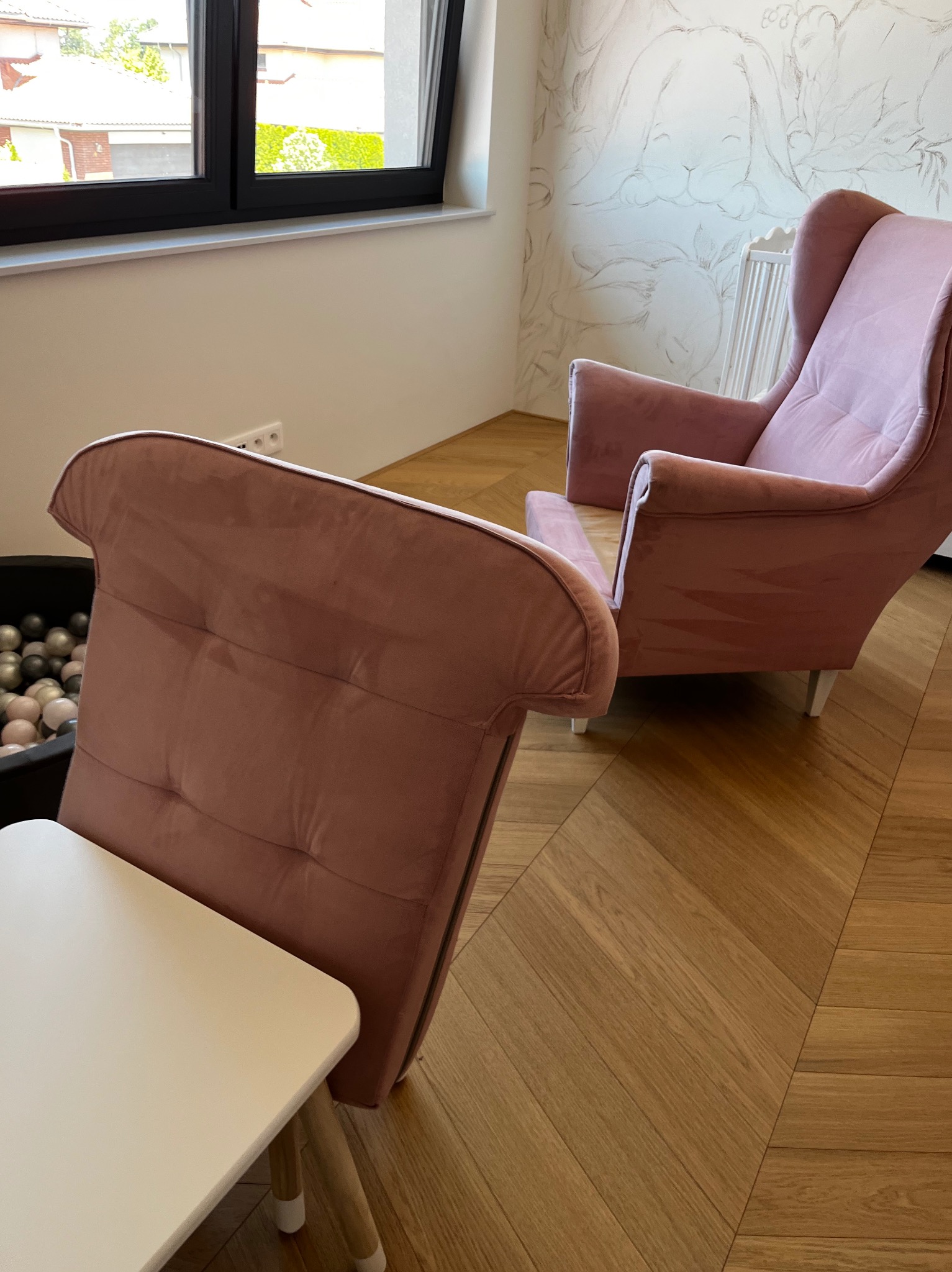 Dwa różowe fotele tapicerowane w pokoju dziecięcym z parkietem w jodełkę, obok stolik i basen z kulkami, na ścianie rysunki zwierząt.
