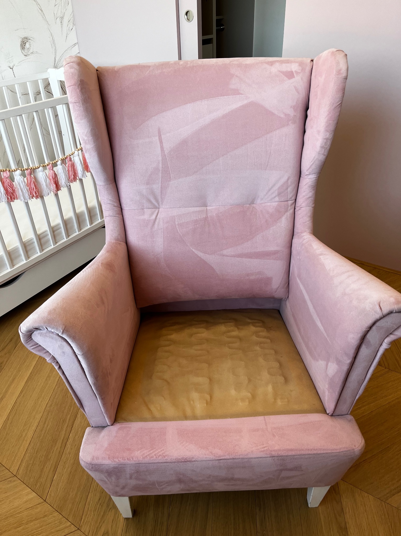 Różowy fotel typu uszak z widocznymi śladami czyszczenia na oparciu i siedzisku, ustawiony w pokoju dziecięcym obok białego łóżeczka ze sznurkiem dekoracyjnym.
