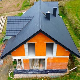 Usługi Budowlane Jania Andrzej - Widok z lotu ptaka na pomarańczowy dom w trakcie budowy z czarnym dachem, widoczne ściany z pustaków, przygotowane drewno i otaczająca zieleń.