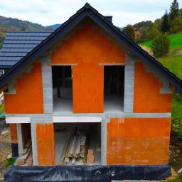 Usługi Budowlane Jania Andrzej - Surowy stan domu z czerwonej cegły z czarnym dachem na tle zielonych wzgórz, widoczne otwory okienne i drzwiowe, składowane drewno przy budynku.