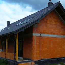 Usługi Budowlane Jania Andrzej - Surowy stan domu jednorodzinnego z czerwonej cegły ceramicznej, z czarną dachówką i drewnianą konstrukcją werandy, widok z zewnątrz na tle pochmurnego nieba.