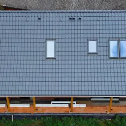 Widok z góry na dom w trakcie budowy z szarą dachówką, trzema oknami dachowymi różnej wielkości i kominem, ściany wzniesione z czerwonej cegły kratówki.