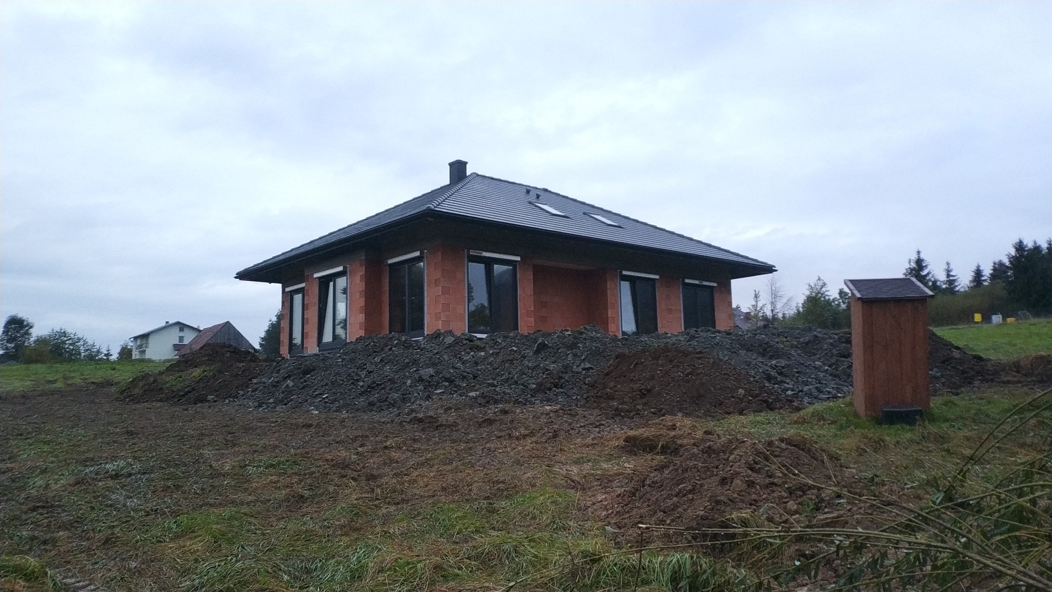 Surowy stan domu murowanego z czarną dachówką i oknami, otoczony stertą ziemi i gruzu, na tle pochmurnego nieba i zieleni.