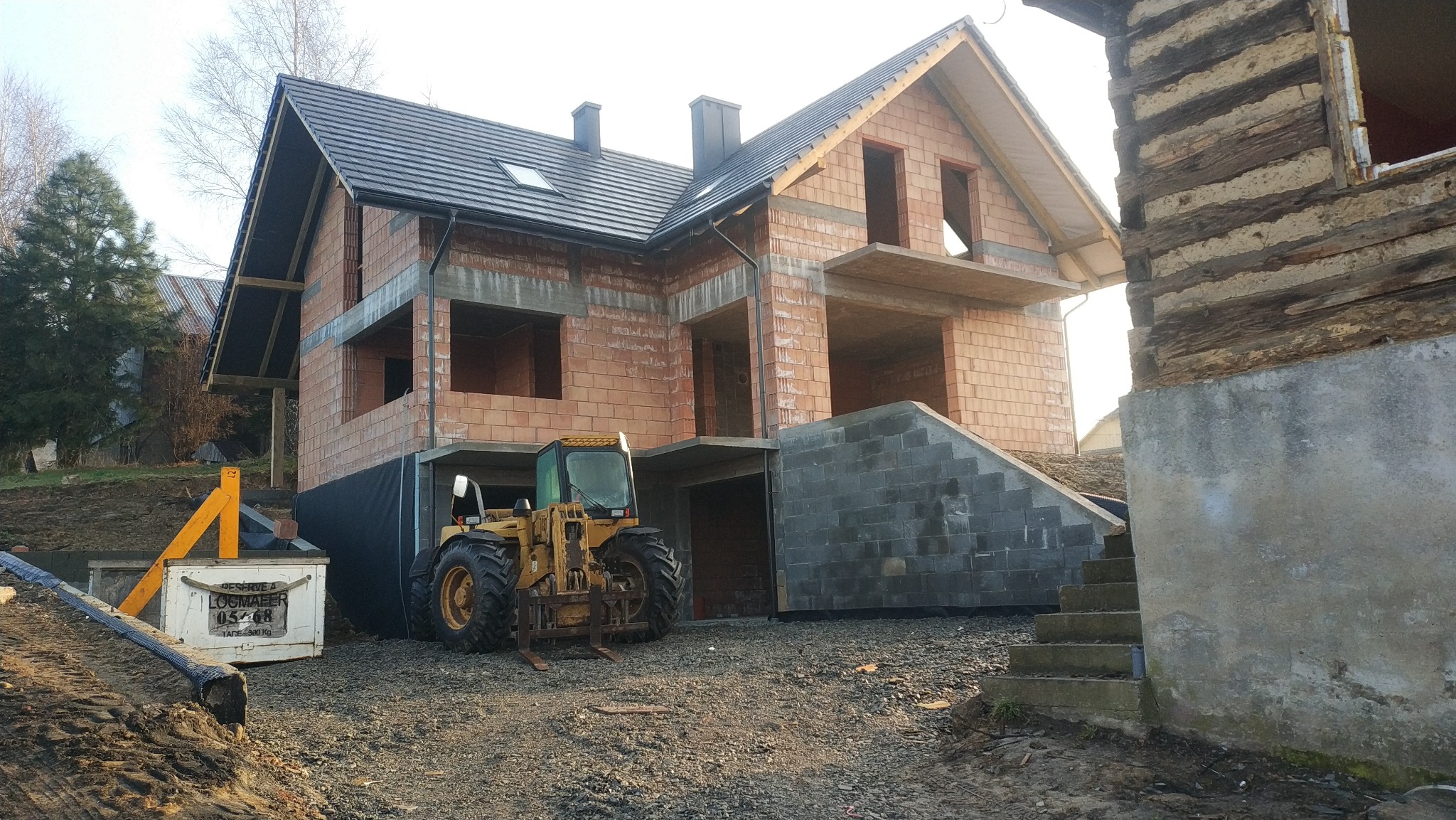 Surowy stan domu murowanego z dachem, widoczna maszyna budowlana i fragment starego drewnianego budynku w tle.
