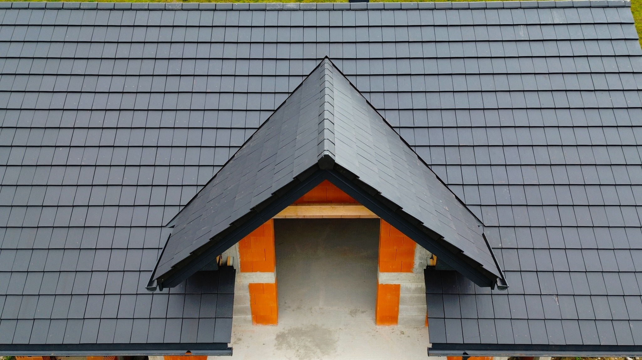 Widok z góry na fragment dachu krytego ciemnoszarą dachówką z wykończoną lukarną w stanie surowym otwartym, z widoczną konstrukcją drewnianą i fragmentami muru z cegły.