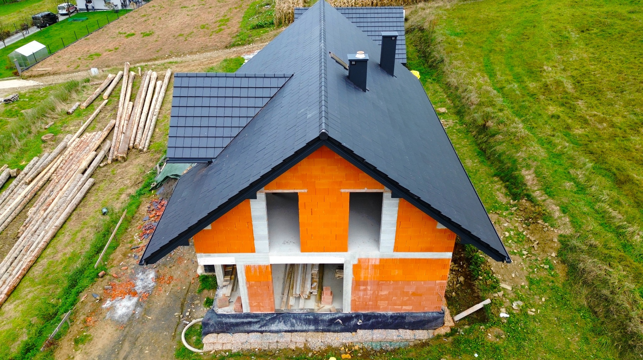 Widok z lotu ptaka na pomarańczowy dom w trakcie budowy z czarnym dachem, widoczne ściany z pustaków, przygotowane drewno i otaczająca zieleń.