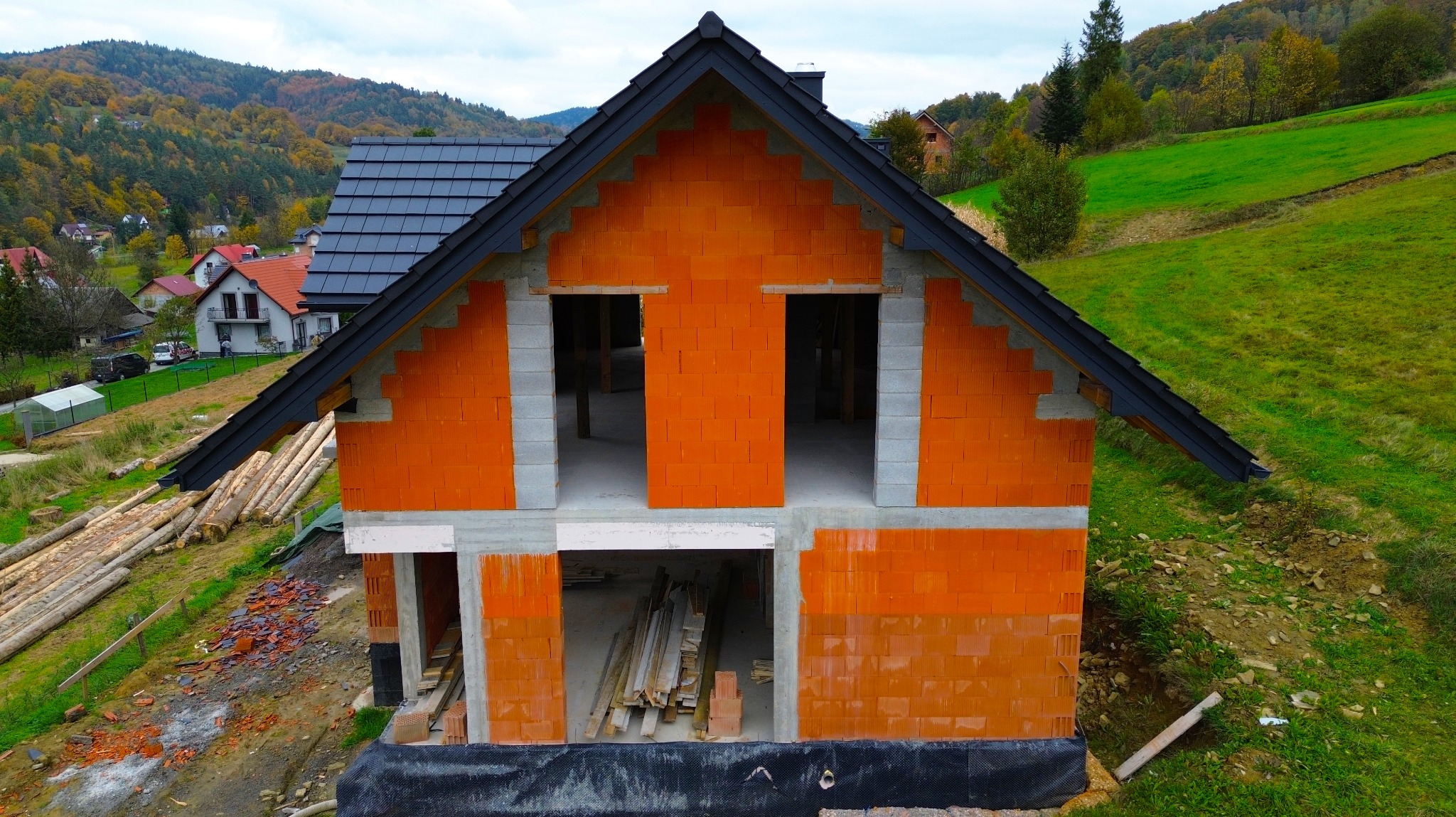 Surowy stan domu z czerwonej cegły z czarnym dachem na tle zielonych wzgórz, widoczne otwory okienne i drzwiowe, składowane drewno przy budynku.